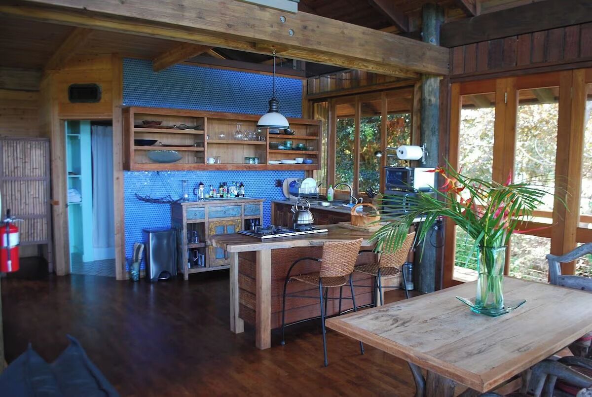 A rustic kitchen with wooden shelves, a central island, and a dining area with a table and chairs.