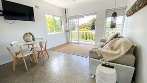 Een moderne woonkamer met een bank, een eettafel met stoelen, een flatscreen televisie aan de wand en grote ramen met uitzicht op de omgeving.