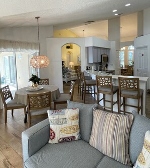 A spacious living area with a gray sofa, wooden chairs, a round dining table, and a kitchen in the background.