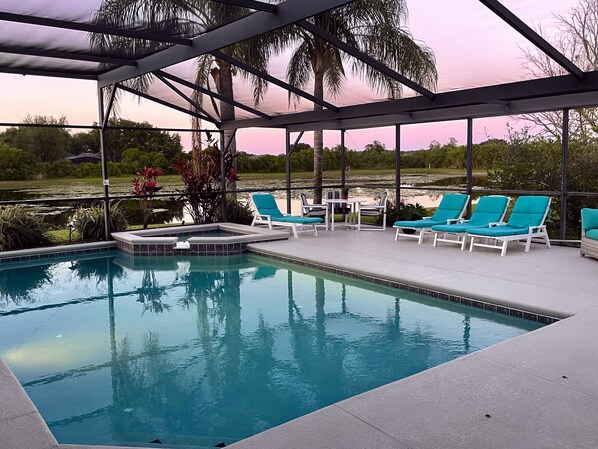 A pool area with lounge chairs and a hot tub, surrounded by a covered patio with a view of a lake and palm trees.