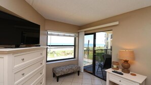 A hotel room with a TV on a dresser, a bench, a desk with a lamp, and a balcony with a view of the ocean.