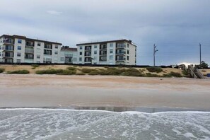 Un edificio frente a la playa con varios pisos, techo plano y una playa arenosa al frente.