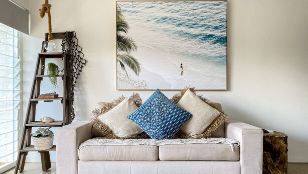 Un salón con un sofá color crema, dos almohadones estampados, un cuadro con una playa, una escalera de madera con plantas y una mesa auxiliar de madera rústica.