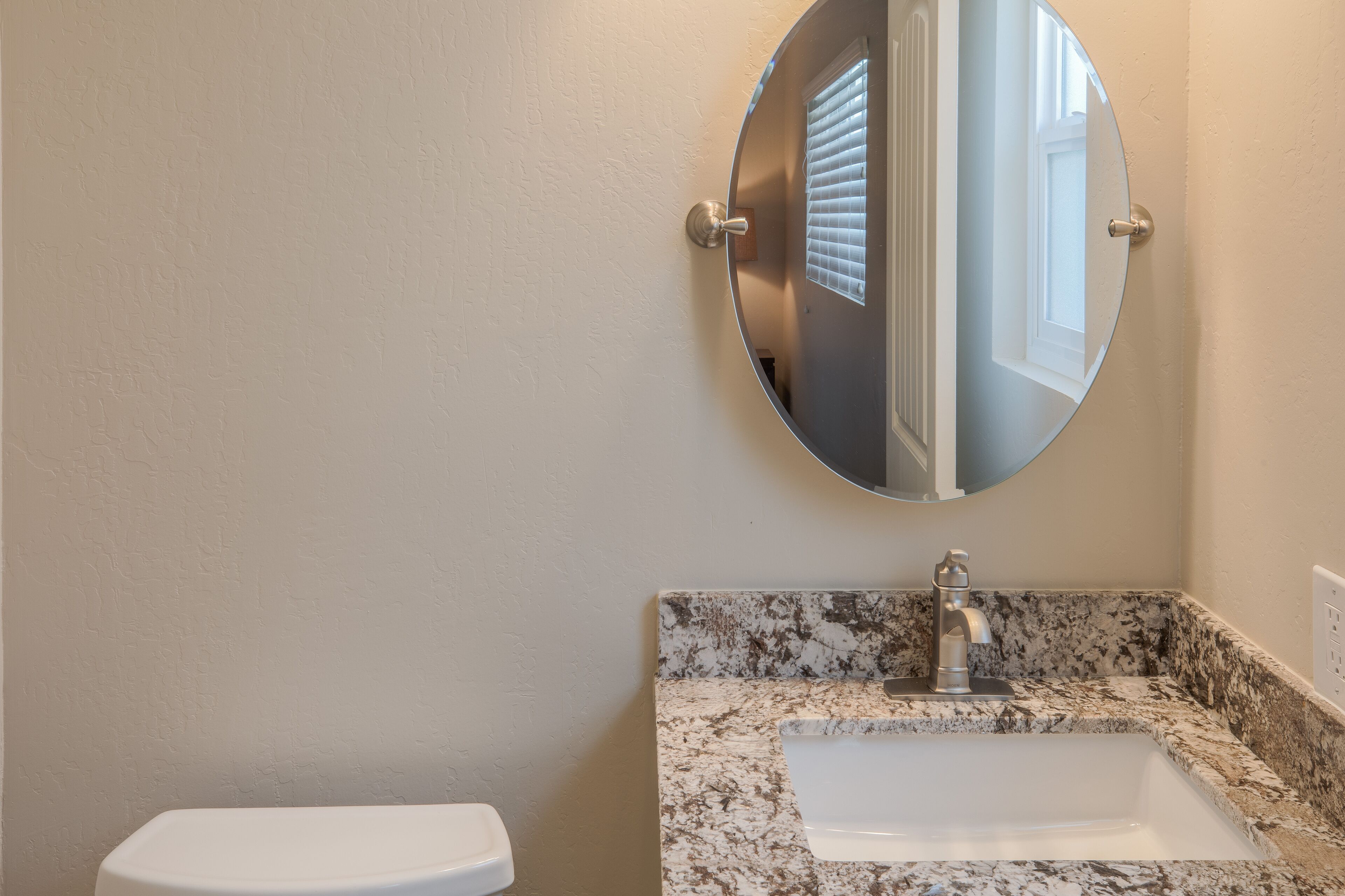 A bathroom with a white toilet, a granite countertop sink, and a round mirror.