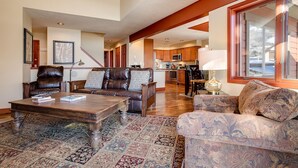 A spacious living room with a leather sofa, patterned armchair, and a coffee table. A kitchen is visible in the background, and there is a large window with a view of the outdoors. - RA208 | Nicely Appointed Condo | Quiet setting | Winter Shuttle | Wood Fireplace (Steamboat Springs)