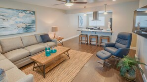 Una sala moderna con un sofá, una mesa de centro y una silla azul. Hay una zona de cocina con taburetes y una mesa de comedor.