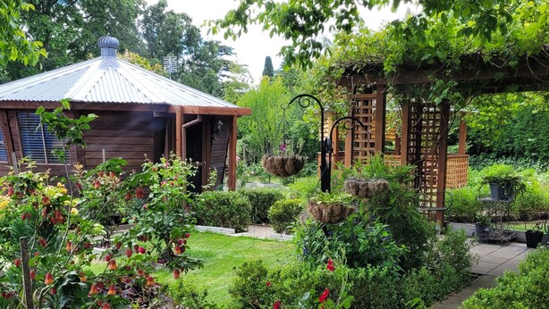 A wooden cabin with a metal roof, surrounded by lush greenery and flowering plants.