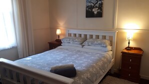 Un dormitorio con una cama blanca, dos mesitas de noche con lámparas, una ventana con cortinas y un cuadro en la pared.