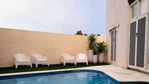 Ein Pool mit einer gefliesten Kante, umgeben von einer beigen Wand und einem Gebäude mit großen Fenstern. In der Nähe des Pools befinden sich drei weiße Kunststoffstühle und zwei Kübelpflanzen.