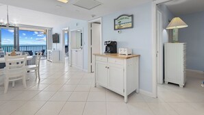 A bright, spacious room with a view of the ocean, a dining area with white furniture, and a kitchen area with a coffee maker and a lamp.