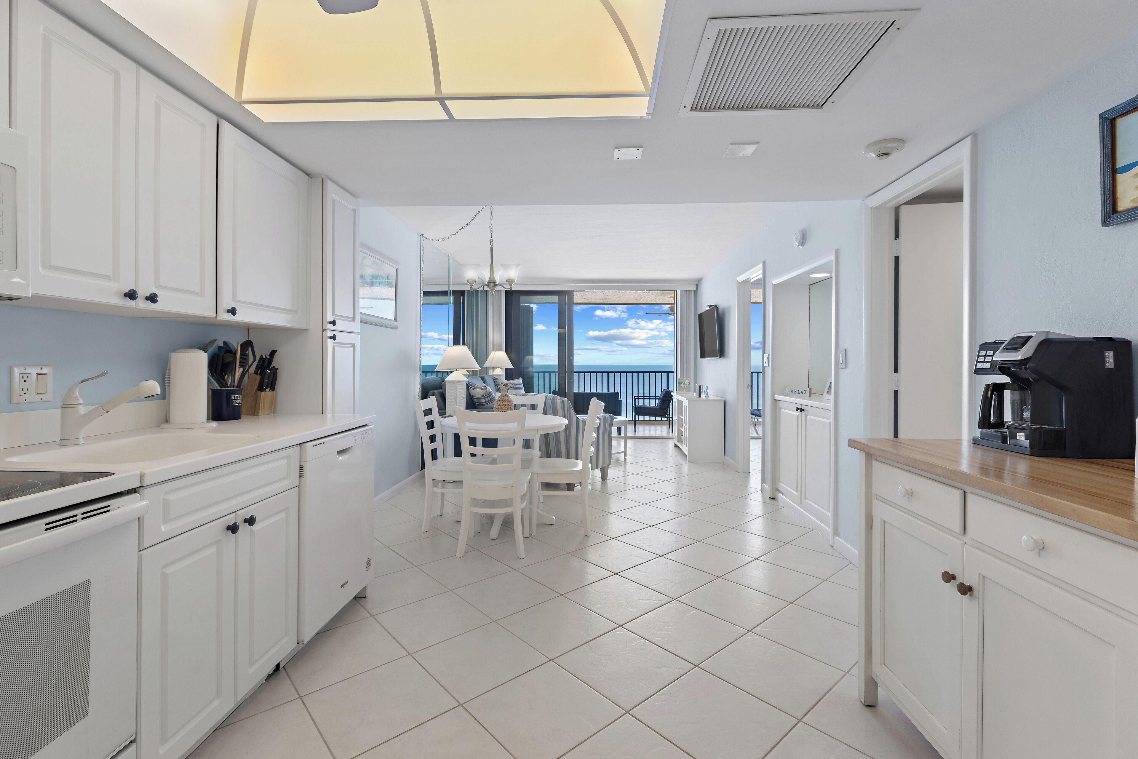 Una cocina moderna con armarios blancos, un comedor con sillas blancas y vistas al mar a través de amplios ventanales.