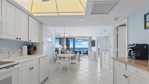 A modern kitchen with white cabinets, a dining area with white chairs, and a view of the sea through large windows.