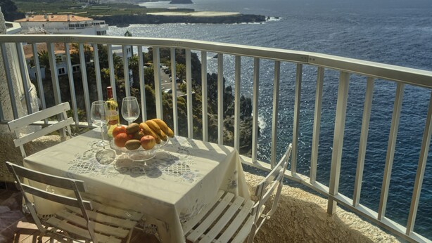 Un balcon avec une table dressée pour deux personnes, offrant une vue imprenable sur la côte, avec des montagnes et la mer en toile de fond.