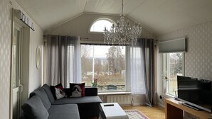 Un salon moderne avec un canapé d’angle, un téléviseur posé sur un meuble en bois, un lustre et une grande fenêtre agrémentée de rideaux.
