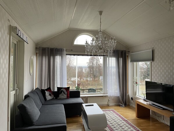 A modern living room with a sectional sofa, a TV on a wooden stand, a chandelier, and a large window with curtains.