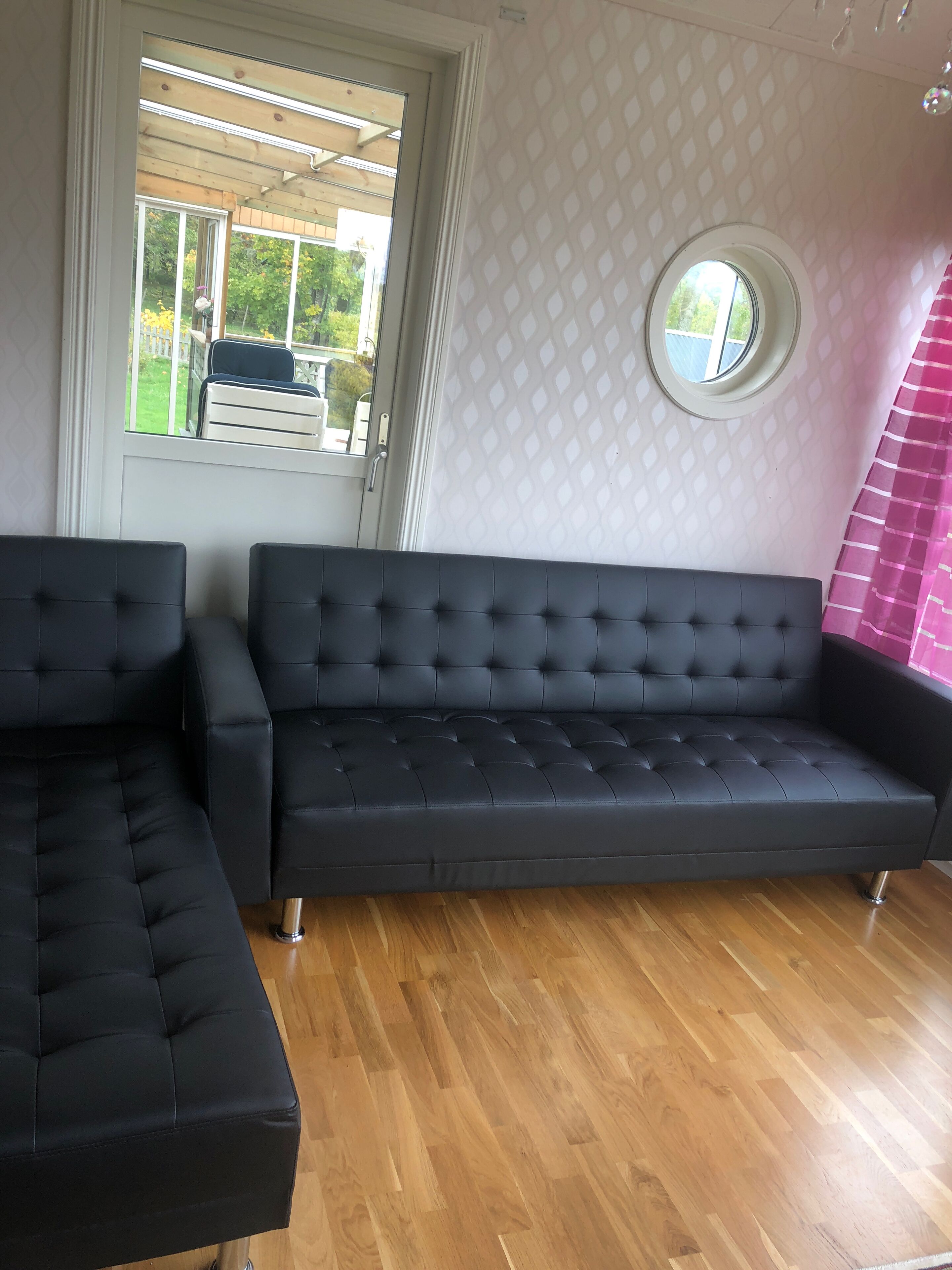 Een kamer met een zwart hoekbank, een houten vloer, een raam met uitzicht op buitenmeubels en een kamer met een patroon op de muur.