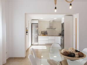 Une cuisine moderne avec des meubles blancs, un réfrigérateur en acier inoxydable et une table à manger en verre sur laquelle est posé un bol rempli de boules décoratives.