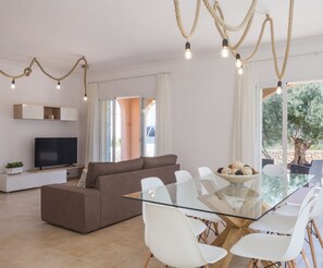 Un salon moderne avec une table à manger en verre, des chaises blanches, un canapé marron et un téléviseur à écran plat.