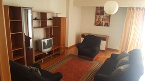 Una sala de estar con un televisor en un estante de madera, un sillón negro, un tapete rojo y una pared con una fotografía de un paisaje urbano.