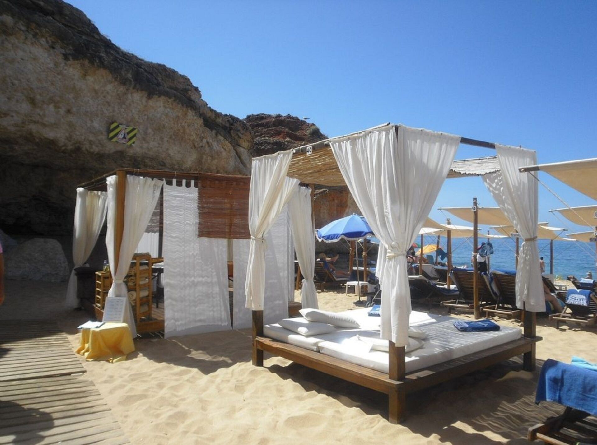 A beach cabana with a wooden bed and white canopy, surrounded by beach chairs and umbrellas.