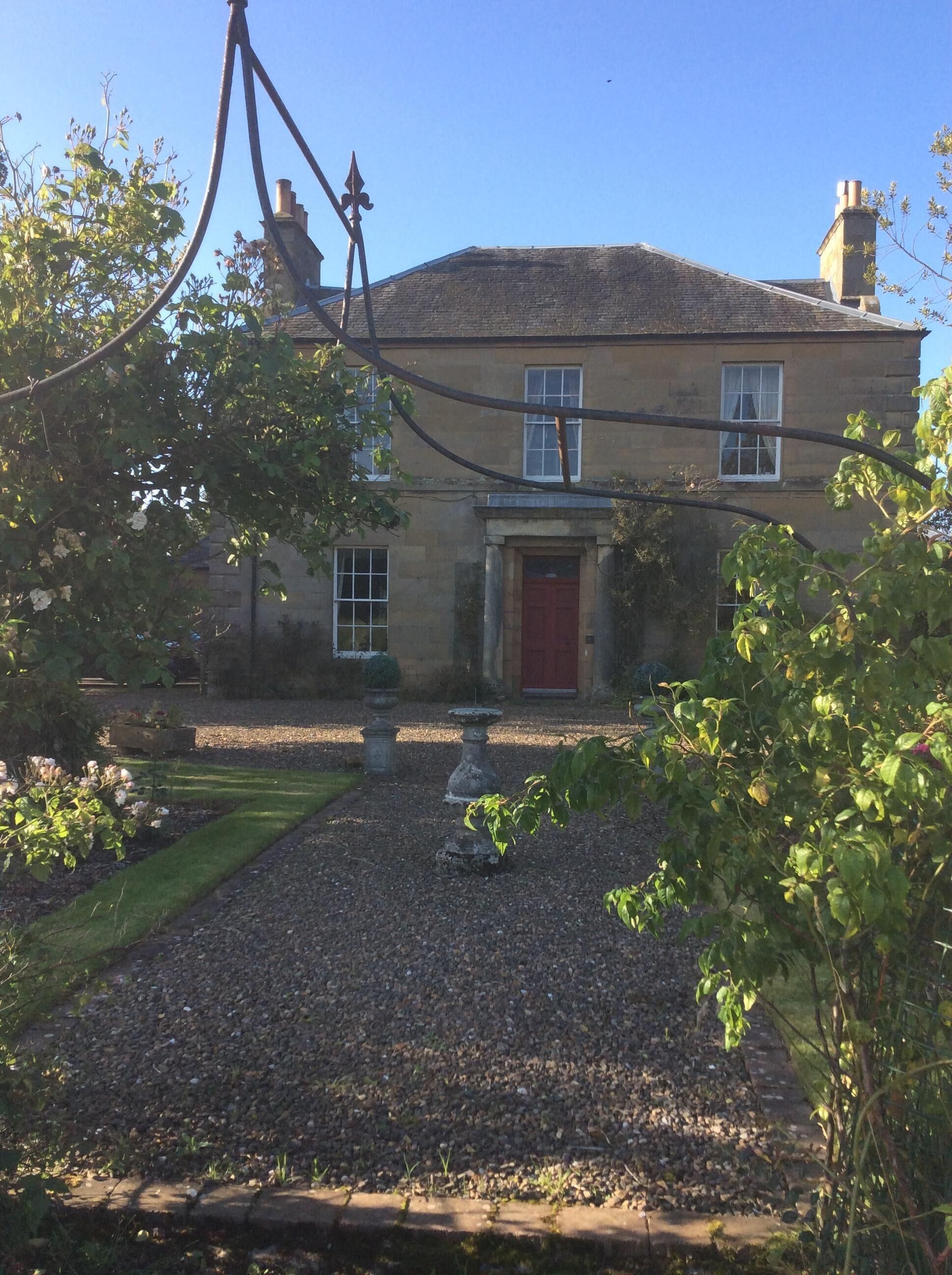 Un bâtiment en pierre avec une porte rouge, un jardin avec des allées de gravier et un ciel bleu clair.