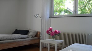 A bedroom with a bed, a bedside table with a vase of flowers, a lamp, and a window with a view of greenery outside. - Quiet non-smoking vacation apartment w beautiful garden in Freiburg-Gundelfingen (Gundelfingen)