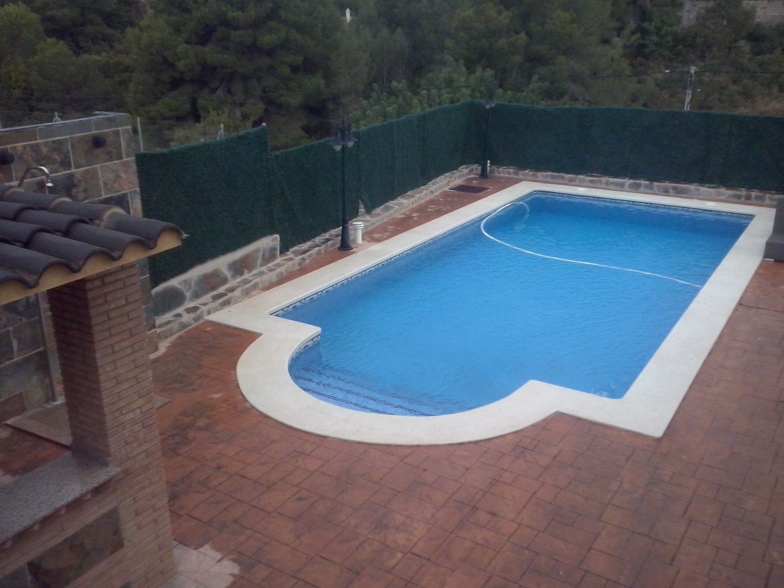 Ein rechteckiger Pool mit geschwungener Kante, umgeben von einer Pflasterterrasse und einem grünen Zaun.