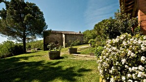 Un bâtiment en pierre, surmonté d’un toit de tuiles, entouré d’une végétation luxuriante et d’arbustes en fleurs.
