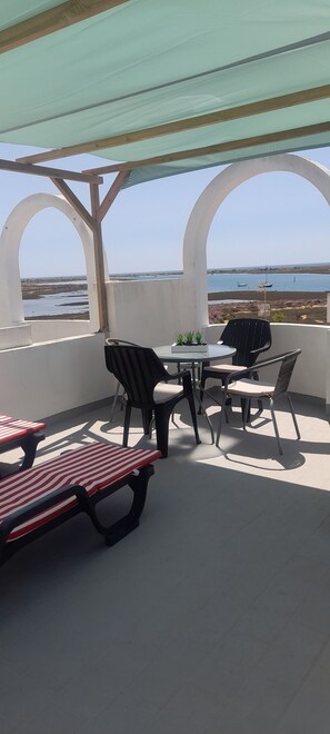 An outdoor patio with a round table and chairs, a picnic table, and a view of the sea.