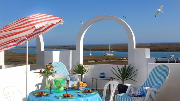 A terrace with a table set for two, a striped umbrella, and a view of the sea.