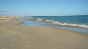 A wide sandy beach with gentle waves, a clear sky, and a person standing in the distance.