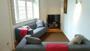 A living room with a grey sofa, a red throw blanket, a television on a wooden stand, and a window with white curtains. - Carolina M Apt in front of Metro (Porto)