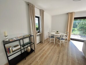 Um quarto com uma mesa de jantar, cadeiras, um console preto com livros e uma vela, e uma janela com cortinas.