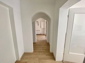 Ein Korridor mit weißen Wänden und Holzboden, der zu einem Raum mit Heizkörper und Tür führt.