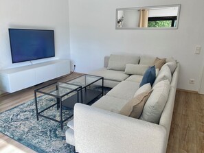 Ein modernes Wohnzimmer mit Ecksofa, Glastisch, Flachbildfernseher und einem großen Spiegel an der Wand.
