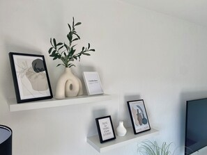 Eine schlichte Wand mit drei Regalen, auf denen gerahmte Kunstwerke, ein Topfpflanze und eine kleine weiße Vase präsentiert werden.