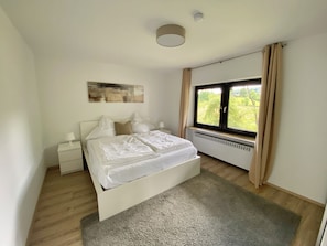 Ein Schlafzimmer mit einem Bett, einem Nachttisch, einer Nachttischlampe, einem Fenster mit Vorhängen und einer Deckenleuchte.