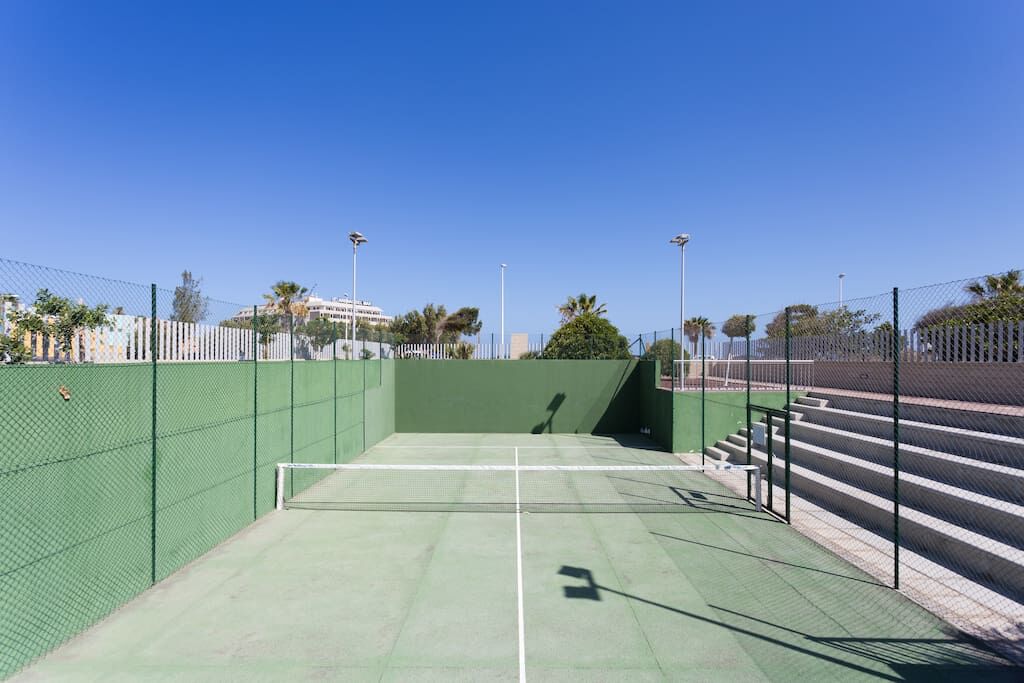 Ein Tennisplatz im Freien mit grüner Belag, umgeben von einem Maschendrahtzaun und blauem Himmel im Hintergrund.