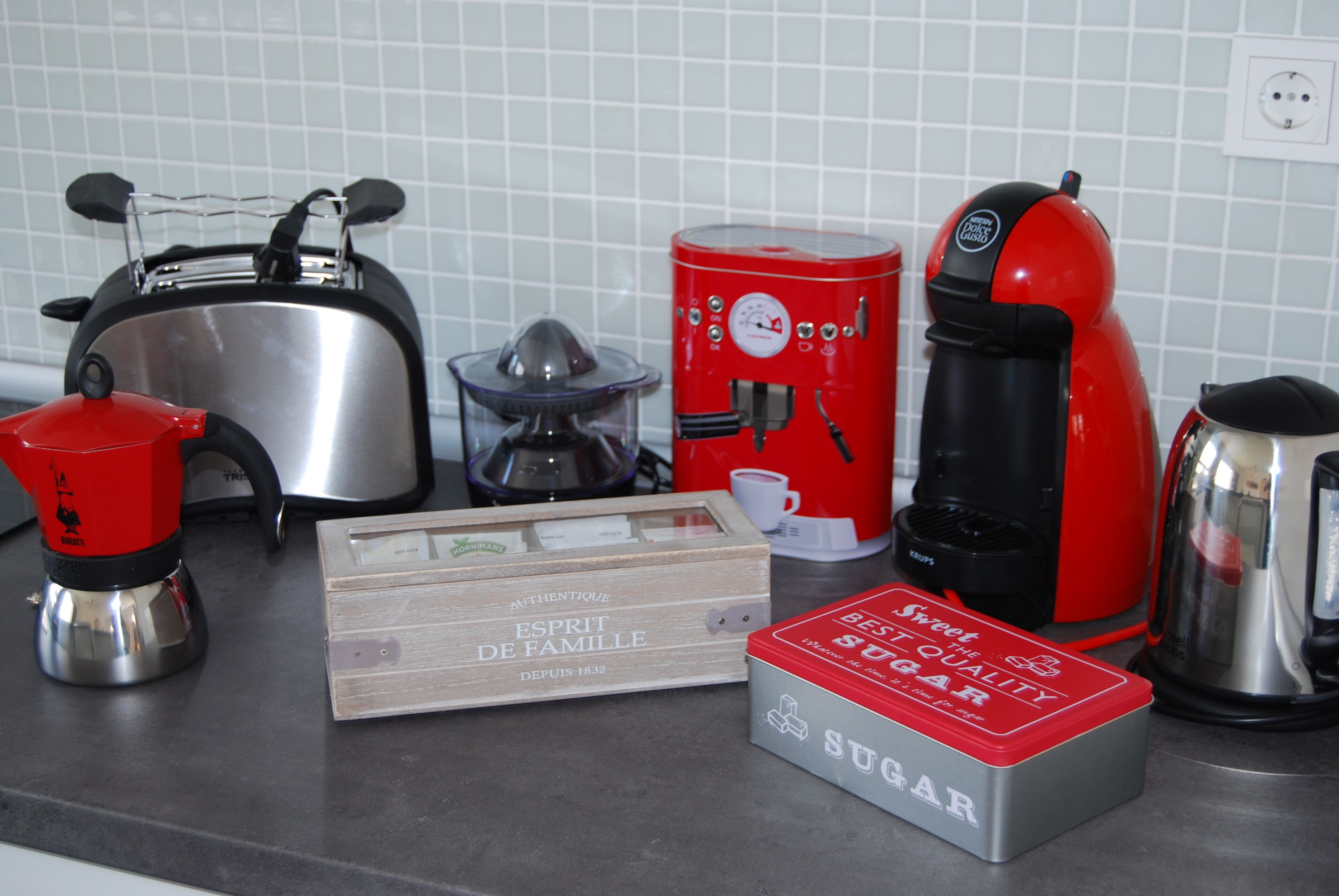 Un plan de travail de cuisine avec un grille-pain, une machine à café, une bouilloire, un récipient à sucre et une boîte en bois étiquetée « Esprit de famille ».