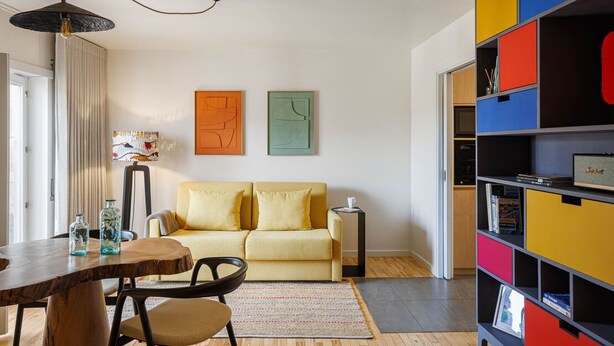 A modern living room with a wooden dining table, yellow sofa, colorful storage unit, and abstract wall art.