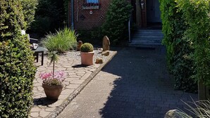 Ein Haus mit geneigtem Dach, einer Veranda und einem Garten mit Kübelpflanzen und einem Weg zum Eingang.