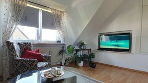 Ein Wohnzimmer mit einem Glastisch, einem gemusterten Sessel, einem roten Kissen, einer Topfpflanze und einem Fernseher an der Wand.