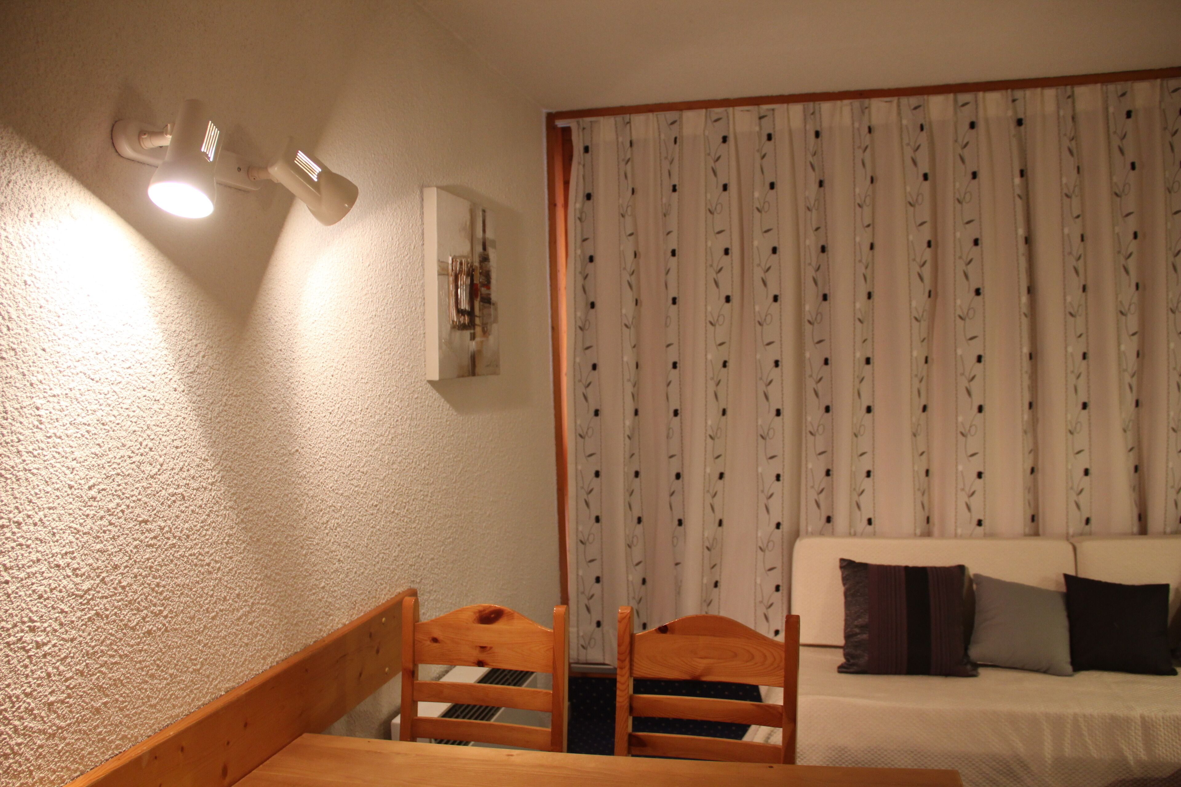 Une pièce comprenant une table et des chaises de salle à manger en bois, un canapé blanc avec des coussins, un rideau à motifs et un luminaire suspendu au plafond.