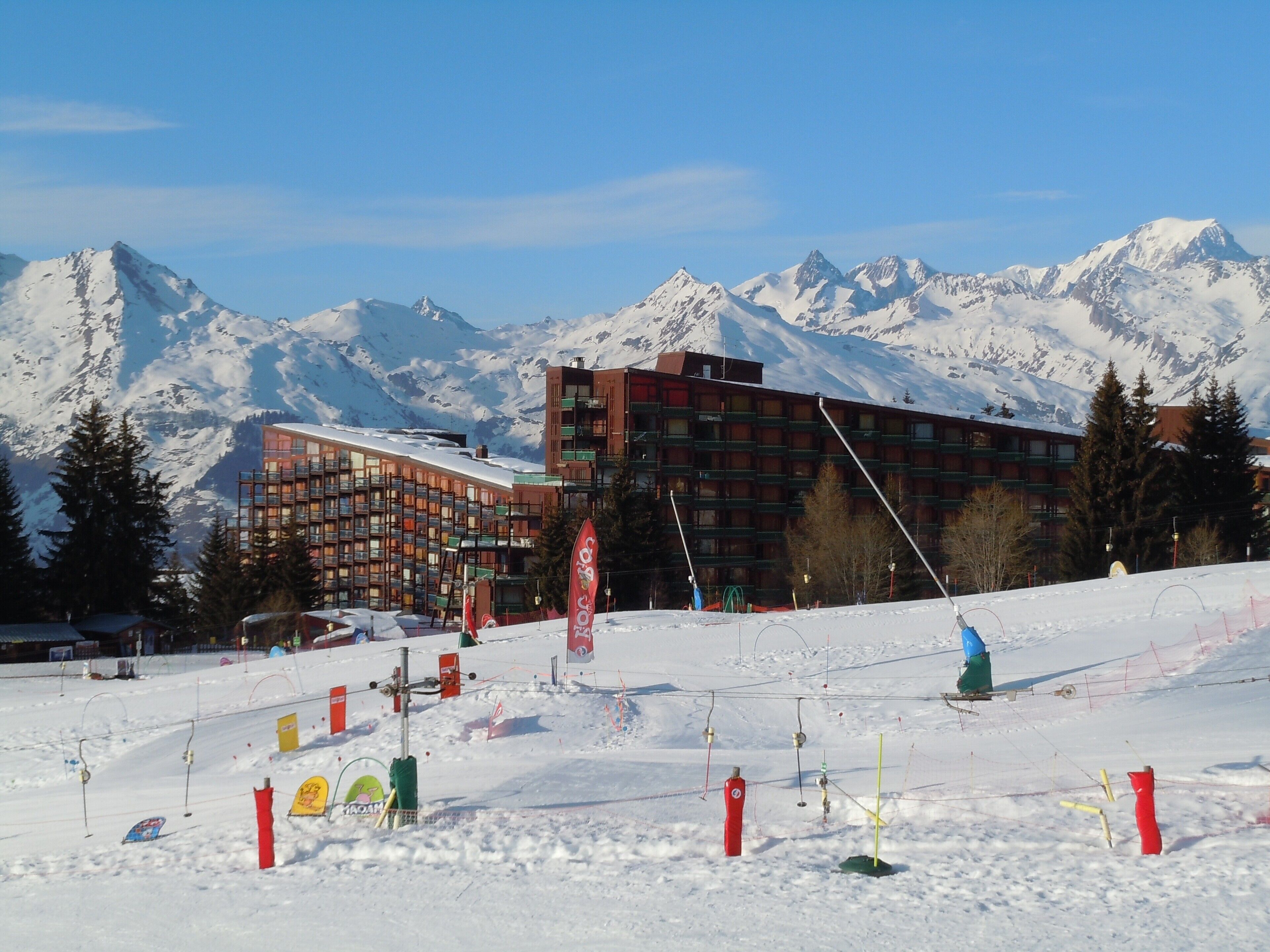 A ski resort with a mountain backdrop, featuring a modern building with multiple floors and balconies, surrounded by snow and trees.