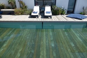 A poolside area with two lounge chairs and a blue cushion, surrounded by plants and a wooden deck.
