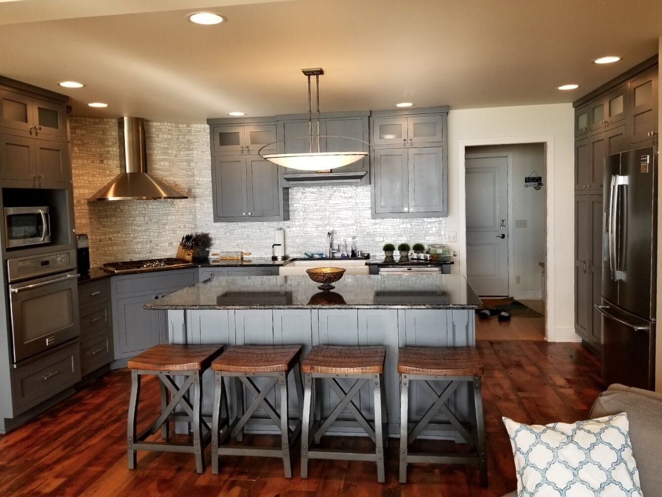 A modern kitchen with grey cabinets, a central island with stools, stainless steel appliances, and a stone backsplash.