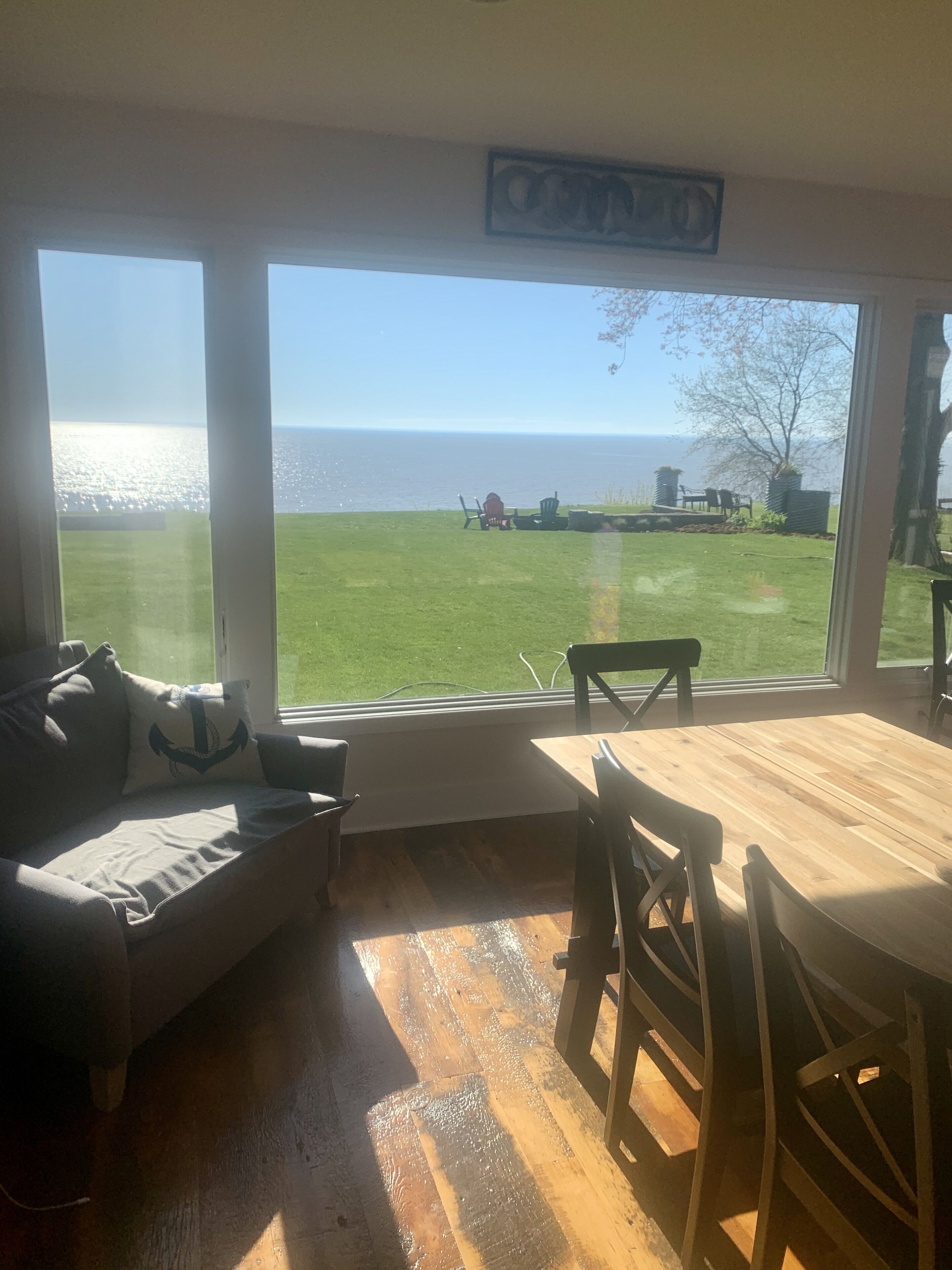 A room with a large window overlooking a grassy area and the sea. There is a wooden table and chairs, a sofa with cushions, and a small rug on the floor.