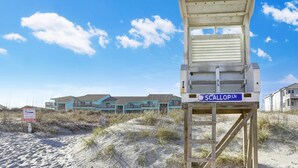 A lifeguard tower labeled "SCALLOP LN" on a sandy beach with dune vegetation, a clear blue sky, and buildings in the background.