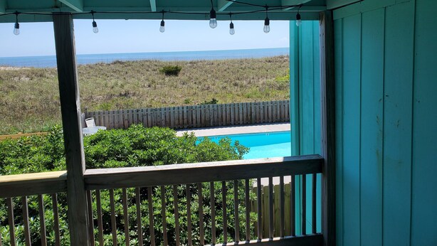 A view from a balcony with a wooden railing, overlooking a swimming pool and a grassy landscape with a fence.