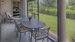 A covered patio with a round table and chairs, a view of a lawn and trees, and a brick wall.